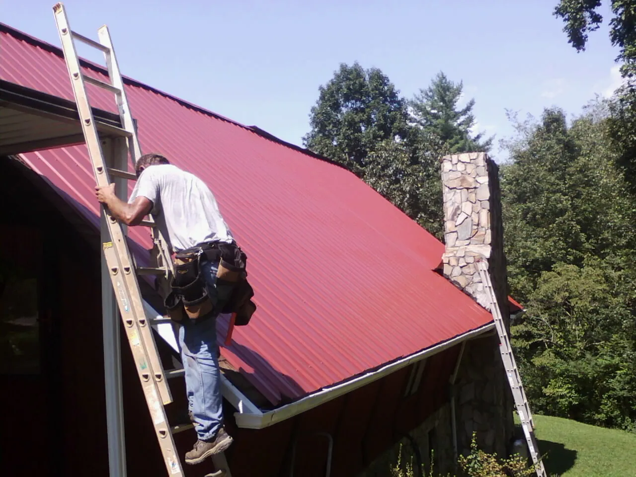 Roofer on a ladder beside a red metal roof and stone chimney — Soffit & Fascia Repair by Roofing Brothers in De Beque